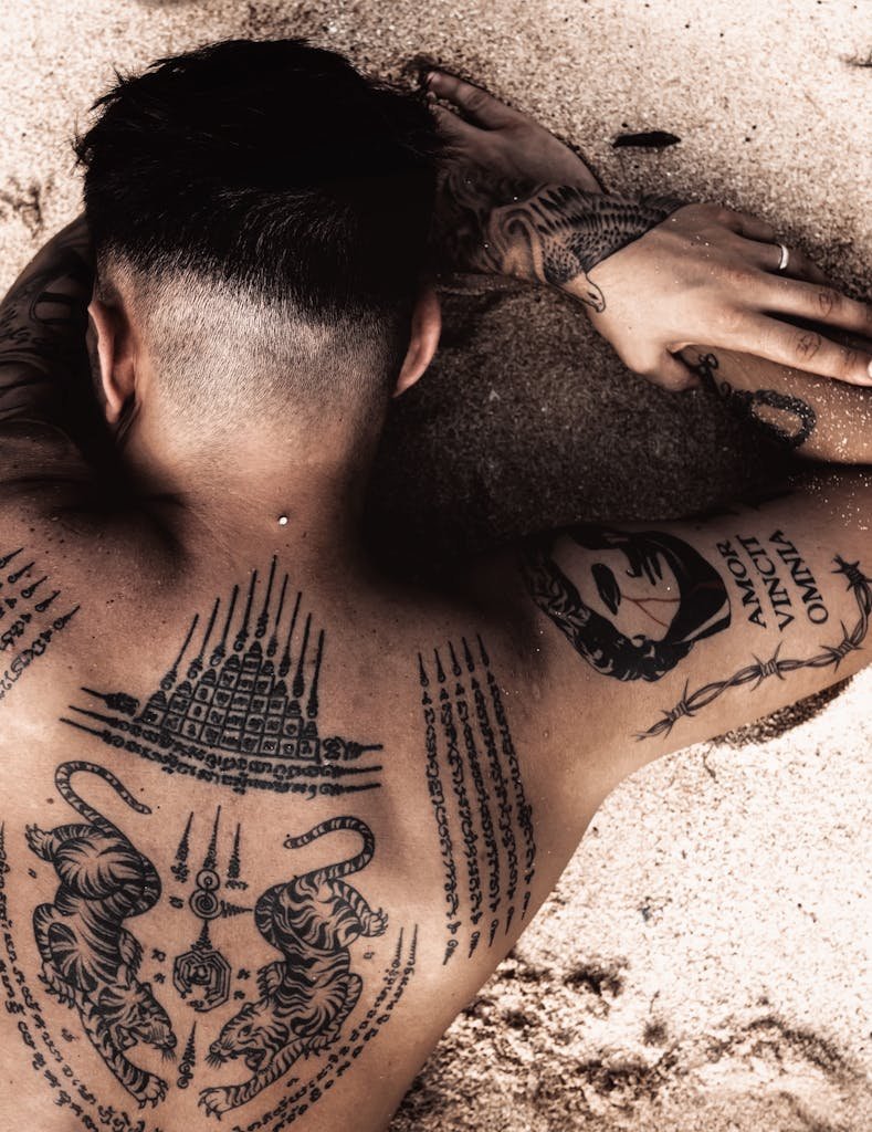 A tattooed man with intricate designs lying on a sandy beach, showcasing body art.